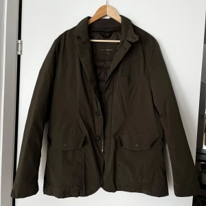 Olivgrön kavajjacka från Massimo Dutti - Säljer en olivgrön kavajjacka från Massimo Dutti med klassisk krage, knappar och två stora fickor framtill. Jackan har quiltat foder på insidan och är perfekt för kyligare dagar. Snygg och stilren design som passar till många tillfällen.
