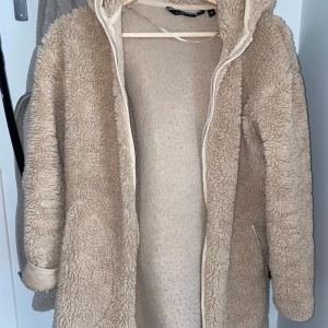 Beige teddyjacka från KappAhl - Mysig beige teddyjacka från KappAhl med huva och dragkedja framtill. Jackan har långa ärmar och två fickor med dragkedja. Perfekt för kyliga dagar och ger en trendig look.
