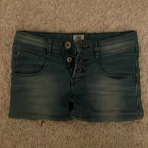 Blå jeansshorts med dubbla knappar - Snygga blå jeansshorts från Tumble ’n dry med dubbla knappar i midjan och klassiska fickor både fram och bak. Shortsen har en lätt tvättad look och bälteshällor för extra stil.