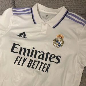 Real Madrid hemmatröja 2022/23 från Adidas - Säljer en vit Real Madrid hemmatröja från Adidas med klubbmärke och sponsortryck på bröstet. Tröjan har korta ärmar, klassisk krage med knappar och lila detaljer på axlarna. Perfekt för fotbollsfans! Köpt på adidas butik i Milano. Ord pris 1100kr