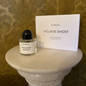 Byredo Mojave Ghost Eau de Parfum - Byredo Mojave Ghost Eau de Parfum i en stilren, genomskinlig glasflaska med svart rund kork. Parfymen levereras med en vit kartongförpackning med minimalistisk design och tydlig logotyp.