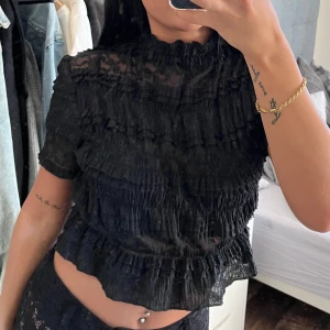 Svart spetts topp  - Säljer en svart transparent blus med volangdetaljer och hög hals. Blusen har korta ärmar och är lätt croppad. Perfekt för dig som gillar trendiga och unika plagg.