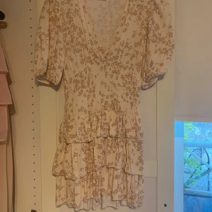 Blommig kortklänning med volang - Söt kortklänning med ljus botten och små beige blommor. Klänningen har v-ringning, puffärmar och flera volanglager i kjolen. Perfekt för sommarens tillfällen och har en feminin, romantisk stil.