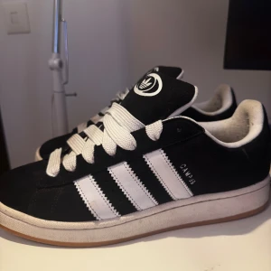 Adidas Campus svartvita sneakers - Säljer ett par klassiska Adidas Campus sneakers i svart mocka med vita ränder och vit sula. Skorna har rund tå, platt sula och breda vita snören. Perfekta för dig som gillar retrostil och streetwear.