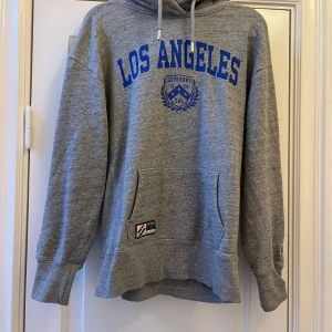 Grå hoodie från Superdry med blått tryck - Säljer en grå hoodie från Superdry med blått 'Los Angeles'-tryck på bröstet och känguruficka framtill. Klassisk huva med dragsko och ribbade muddar. Perfekt för en avslappnad stil.