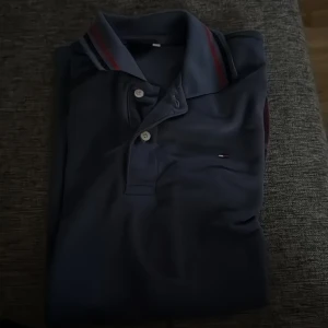 Mörkblå piké t-shirt från Tommy Hilfiger - Snygg mörkblå piké t-shirt från Tommy Hilfiger med klassisk krage och knappar framtill. Kragen har röd och vit randig detalj och en liten logga på bröstet. Perfekt för en stilren look.