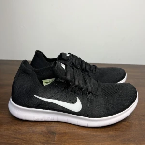Svarta Nike Free Flyknit springskor - Svarta Nike Free Flyknit sneakers med vit sula och vit Nike-logga på sidan. Skorna har snörning och är tillverkade i ett stickat material som ger en lätt och flexibel känsla. Perfekta för träning eller vardagsbruk. Funkar som springskor och inomhus!