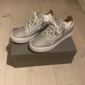 Glittriga sneakers från Giuseppe Zanotti - Säljer ett par riktigt snygga sneakers från Giuseppe Zanotti i silver med glittrig finish. Skorna har vita snören, dragkedjor på sidorna och rund tå. Perfekta för dig som vill sticka ut med din stil!