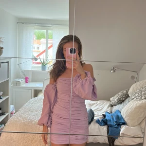 Ljuslila offshoulder klänning från Gina Tricot - Supersöt ljuslila kortklänning från Gina Tricot med offshoulder och långa puffärmar. Klänningen har rynkade detaljer framtill och en figurnära passform som framhäver siluetten. Perfekt för dig som vill sticka ut med en trendig look!