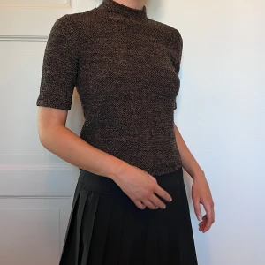 Brun och glittrig stickad polotopp - Säljer en brunmelerad stickad polotopp med korta ärmar och hög hals. Toppens material har en tydlig struktur. Storlek M. 