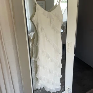 Vit långklänning med volanger - Säljer en vit långklänning med smala axelband och volangdetaljer som löper diagonalt över hela klänningen. Använd på midsommar 1 gång. Storlek M 