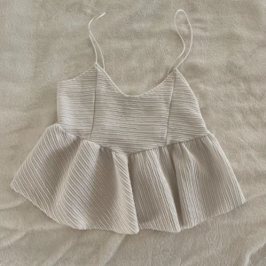 Vit plisserad linnetopp från Zara - Säljer en vit plisserad linnetopp från Zara med tunna axelband och v-ringad hals. Toppen har en lös passform och volang nedtill, vilket ger en luftig och trendig look. Perfekt till varma dagar eller lager på lager.