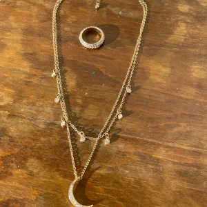 Guldfärgat halsband och ring med stenar - Säljer ett set med ett guldfärgat halsband med dubbla kedjor, små gnistrande stenar och en måne som hänge samt en matchande ring med stenar runt om. Perfekt för dig som gillar detaljer och glittriga accessoarer.