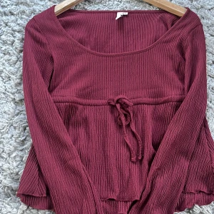 Vinröd långärmad topp från Nelly - Säljer en vinröd långärmad topp från Nelly med knyt i midjan och lätt rynkad struktur. Toppen har rundad halsringning och vida ärmar med volangkant. 