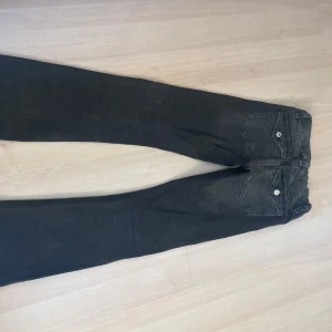Svarta bootcut jeans - Säljer ett par svarta bootcut jeans med låg midja och klassiska fem fickor. Jeansen har vita knappar och en lätt tvättad look. 