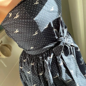 Mörkblå axelbandslös klänning med prickigt mönster från Hollister - Säljer en söt kort  mörkblå axelbandslös klänning från Hollister med vita prickar och små fåglar. Klänningen har en markerad midja med knytband och en lätt utställd kjol. Perfekt för varma dagar! Det är stl M i klänningen men passar även S,