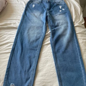 Blå wide leg jeans med slitningar - Säljer ett par blå jeans med wide passform och slitna detaljer framtill. Jeansen har klassisk femficksdesign, knapp och dragkedja i gylfen. Perfekta för en avslappnad och trendig look.