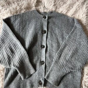 Grå ribbstickad kofta med knappar - Mysig grå kofta med ribbstickat mönster och stora svarta knappar framtill. Koftan har rund halsringning och långa ärmar, perfekt för lager-på-lager-stil.
