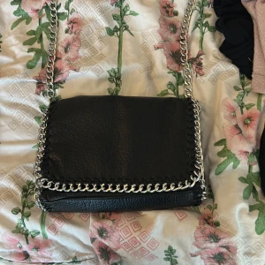 En fin Tiamo väska med axelrem  - Säljer en svart axelväska från Tiamo med silverfärgad kedja som axelrem och runt kanterna. Väskan har dragkedja på baksidan och är tillverkad i syntetmaterial med en cool, stilren look.