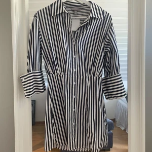 Randig skjortklänning - Helt oanvänd med prislapp kvar! Snygg skjortklänning med svarta och vita vertikala ränder. Klänningen har knappar framtill, krage och långa ärmar med breda manschetter. Perfekt för en stilren och trendig look. 