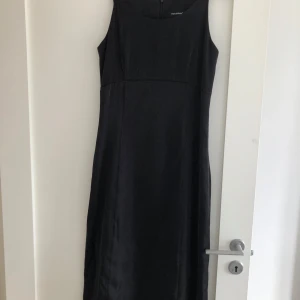 Svart långklänning från Mari Philippe - En stilren svart långklänning från Mari Philippe med ärmlös design och rundad halsringning. Klänningen har en klassisk, figurnära passform och dragkedja i ryggen. Perfekt för dig som gillar enkel och tidlös stil.