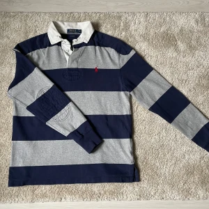 Långärmad Polo Ralph Lauren Piké - Säljer en klassisk rugbytröja från Polo Ralph Lauren i marinblått och grått med breda ränder. Tröjan har några pytte hål men annars u fint skick. Den är i storlek Small och sitter åt slim hållet. Pris kan diskuteras, kontakta gärna vid funderingar!😀