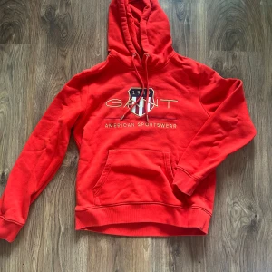 Röd hoodie från GANT - Säljer en röd hoodie från GANT med broderad logga och texten 'American Sportswear' på bröstet. Tröjan har luva och en stor magficka. Den är lite mindre skrikröd än vad den ser ut som på bilden. Perfekt för en sportig och avslappnad stil! 💓