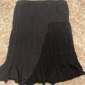 Svart smockad kjol från Gina Tricot - Svart kjol från Gina Tricot i smockat material med lätt volang nertill. Kjolen är elastisk och har en bekväm passform som sitter snyggt på kroppen. Perfekt att styla med en enkel topp eller stickad tröja.