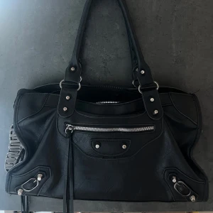 Svart handväska med silverdetaljer - Säljer en svart handväska med silverfärgade metalldetaljer och dragkedjor. Väskan har dubbla handtag, dekorativa spännen och en framficka med dragkedja. Rymligt huvudfack med innerficka. Perfekt för dig som vill ha en stilren och praktisk accessoar.