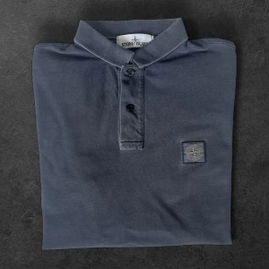 Mörkgrå piké från Stone Island - Säljer en mörkgrå pikétröja från Stone Island med klassisk krage och två knappar framtill. Tröjan har korta ärmar och en broderad logotyp på bröstet. Perfekt för en stilren och avslappnad look.