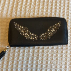 Svart clutchväska/plånbok med nitar - Cool svart plånbok med silvriga nitar i form av vingar på framsidan. Plånboken har dragkedja runtom och en tofs som detalj på dragkedjan. Flera fack för kort och myntfack med dragkedja inuti. Perfekt accessoar för dig som gillar unika detaljer.