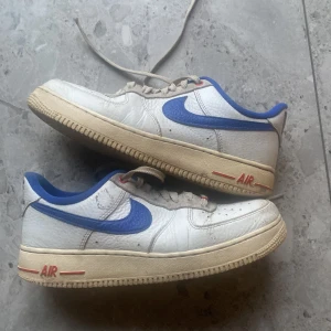 Nike Air Force 1 med blå swoosh - Klassiska Nike Air Force 1 sneakers i vitt skinn med blå swoosh och blå detaljer runt häl och plös. Skorna har beige sula med röd AIR-text på sidan och perforerad tå för ventilation. Låga och med snörning.