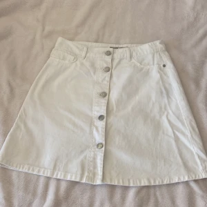 Vit jeanskjol med knappar från Noisy May - Säljer en vit jeanskjol från Noisy May i A-linjeformad modell. Kjolen har knappar framtill, fickor både fram och bak samt hällor för skärp. Perfekt för dig som gillar en enkel och stilren look.