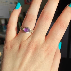 Silverring med lila sten - Säljer en snygg ring i silverfärgad metall med en oval lila sten i mitten. Ringen har en unik tvinnad design runt stenen som ger ett bohemiskt intryck. Perfekt för dig som vill ha något unikt och färgglatt till din smyckessamling.
