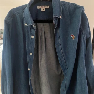 Blå jeansskjorta från U.S. Polo Assn. - Säljer en klassisk blå jeansskjorta från U.S. Polo Assn. med knappar framtill och broderad logga på bröstet. Skjortan har lång ärm och är perfekt för en avslappnad men ändå stilren look.