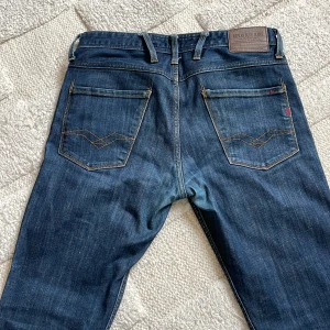 Replay jeans - Säljer ett par Replay Anbass jeans i klassisk blå denim med snygga kontrastsömmar och raka ben. Byxorna har fem fickor, Replay-logga vid fickan och slim-fit passform. Perfekta för dig som gillar stilrena jeans med bra kvalitet.