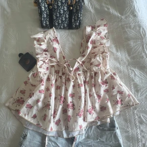 Blommig volangtopp med knytband - Supersöt blommig topp med volangdetaljer och knytning framtill. Toppen är i ljusbeige med rosa blommor och har en luftig, flowy passform. Perfekt för varma dagar och ger en romantisk vibe.