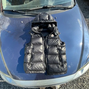 Svart dunväst från Moncler - Säljer en svart dunväst från Moncler med huva och två dragkedjefickor framtill. Västen har Moncler-logga på bröstet och klassisk quiltad design. Perfekt för lager-på-lager under kyliga dagar.