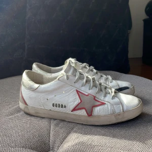 Golden Goose Superstar  - Golden Goose Superstar sneakers i vitt skinn med en röd stjärna på sidan och silverfärgade detaljer vid hälen. Skorna har snörning och platt sula. Nypris runt 6000kr jag säljer för 1299.