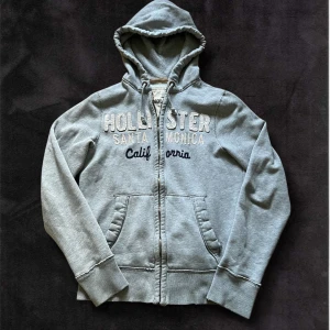 Grå hoodie från Hollister med dragkedja - Grå hoodie från Hollister med broderad text 'Santa Monica California' på bröstet. Tröjan har huva med dragsko, fickor framtill och dragkedja. Mjuk insida och ribbade muddar vid ärmslut och nederkant.