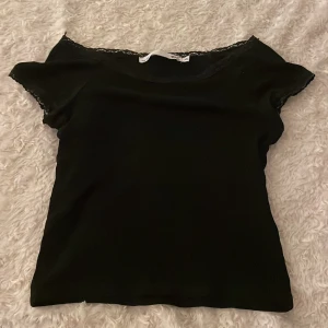 Svart topp med spetsdetalj - Säljer en svart topp med korta ärmar och fin spetsdetalj längs halsringningen. 