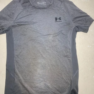 Grå träningst-shirt från Under Armour - Säljer en grå kortärmad träningst-shirt från Under Armour med svart logga på bröstet. Tillverkad i ett syntetmaterial som andas, perfekt för träning och sport. Snygg passform och bekväm att ha på sig.