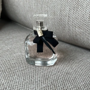 Yves Saint Laurent Mon Paris Eau de Parfum - Elegant parfymflaska från Yves Saint Laurent, Mon Paris. Flaskan är genomskinlig med en fyrkantig form och dekorerad med en svart rosett och gulddetalj vid halsen. Perfekt för dig som gillar lyxiga och feminina dofter. Testad, 50ml flaska 