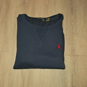 Mörkblå sweatshirt från Polo Ralph Lauren - Klassisk mörkblå sweatshirt från Polo Ralph Lauren med rund halsringning och röd broderad logga på bröstet. Tröjan har långa ärmar och ribbade muddar vid ärmslut och nederkant. Perfekt för en stilren och avslappnad look.