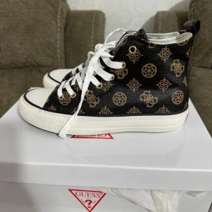 Svarta sneakers med mönster från Guess - Snygga svarta höga sneakers från Guess med bruna logomönster och vita snören. Skorna har vit sula och klassisk rund tå. 