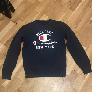 Mörkblå sweatshirt från Champion - Mörkblå sweatshirt från Champion med vit och röd logga samt texten 'ATHL. DEPT. Champion NEW YORK' på bröstet. Klassisk rund halsringning och långa ärmar. Perfekt för en sportig och avslappnad stil. Storlek Small.