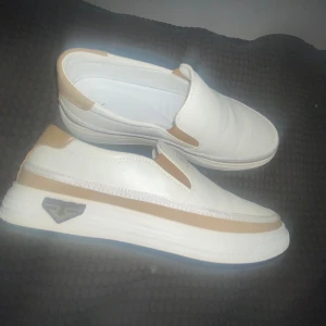 Vita slip-on sneakers med beige detaljer - Säljer ett par fräscha vita slip-on sneakers med beige detaljer och tjock vit sula. Skorna har en stilren design utan snörning och är perfekta för en clean och modern look.