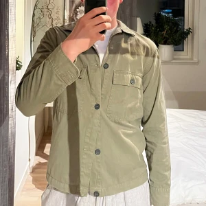 Olivgrön Overshirt - Säljer en olivgrön Overshirt från 157 med klassisk krage och två bröstfickor med knappar. Skjortan har långa ärmar och stängs med knappar framtill. Perfekt för en avslappnad och stilren look. Storlek S men skulle säga att den även skulle passa M.
