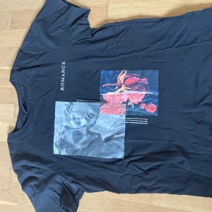 Svart t-shirt med tryck från Gina Tricot - Svart t-shirt från Gina Tricot med coolt tryck framtill som visar en staty och blommor samt texten 'ROMANCE'. T-shirten har rund hals och korta ärmar. Perfekt för en trendig och avslappnad stil.