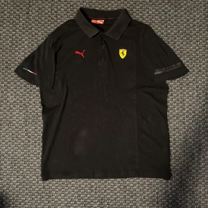 Svart Ferrari x Puma piké t-shirt - Säljer en svart piké t-shirt från Puma i samarbete med Ferrari. T-shirten har klassisk krage, broderad Puma-logga, Ferrari-märke på bröstet och 'Scuderia Ferrari' tryck på ärmen. Italienska flaggan broderad på ena ärmen.